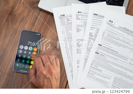 Close-up of a person calculating taxes on a smartphone with tax forms spread out on a wooden desk. Tax preparation. Perfect for accounting, tax season, and financial management Close-up of a person calculating taxes on a smartphone with tax forms spread out on a wooden desk. Tax preparation. Perfect for accounting, tax season, and financial management 123424799
