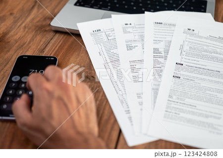 Hand using a smartphone calculator next to various tax forms on a wooden desk. Tax season, accounting, and financial planning concept  123424808