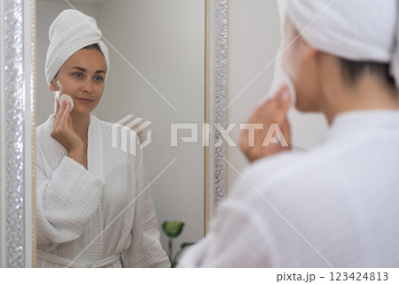 In a bathrobe, a woman enhances her radiance, applying skincare products and captivated by her reflection in the mirror. In a bathrobe, a woman enhances her radiance, applying skincare products and captivated by her reflection in the mirror. 123424813