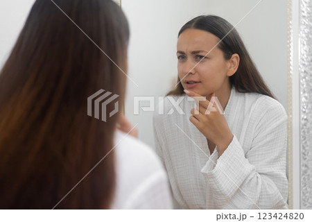In the bathroom, a concerned woman looks at her reflection with sadness, distressed by her skin problems. In the bathroom, a concerned woman looks at her reflection with sadness, distressed by her skin problems. 123424820