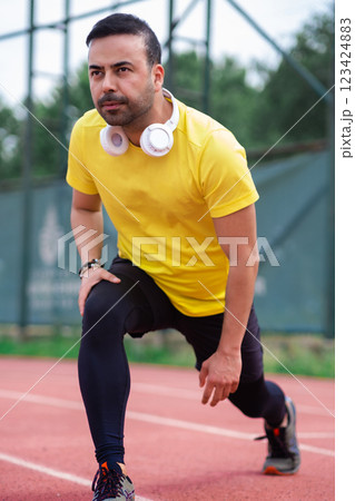 Active sports enthusiast in wireless headphones prepares leg muscles for training on rubberized running track at city park.  123424883