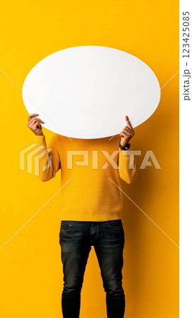 Man wearing yellow sweater and jeans holding large blank white speech bubble in front of face against yellow background, suggesting questions, messaging, and communication 123425085