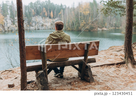 Lonely 30s blonde man is sitting on the bench of a forest lake. Autumn landscape, calmness, tranquility, travel concept. A man on the bank of a lake is enjoying the nature. High quality photo. 123430451