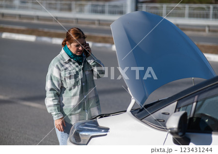 Caucasian woman cries and calls on the phone standing near the car with the hood open.  123431244