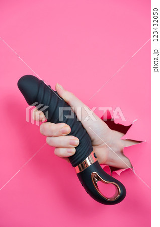 Female hand sticking out of pink paper background with black textured vibrator. Vertical photo.  123432050