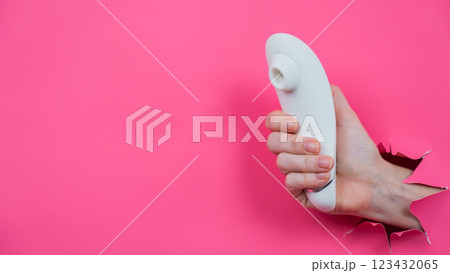 Female hand sticks out white vacuum clitoral vibrator through pink paper background.  123432065