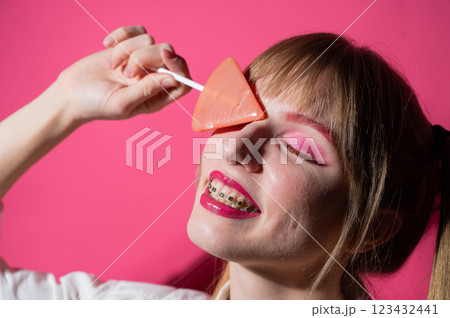Portrait of a young woman with braces and bright makeup eating a lollipop on a pink background. Portrait of a young woman with braces and bright makeup eating a lollipop on a pink background. 123432441