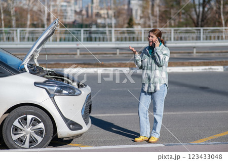 Caucasian woman cries and calls on the phone standing near the car with the hood open.  123433048