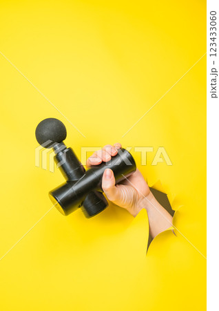 Female hand sticking out with massage gun from yellow paper background. Vertical photo.  123433060