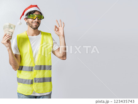 Engineer in christmas hat holding money in ok gesture. builder engineer in christmas hat with approving expression holding money bills. Christmas engineer concept holding money in happy holidays 123435743