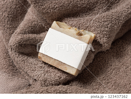 Beige herbal soap bar with blank label on light brown bath towel top view, hygiene product mockup 123437162