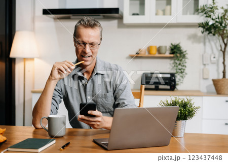 Lifestyle people, using laptop computer and smartphone sitting in the kitchen. Happy mature male, entrepreneur, small business owner working online at home office. Lifestyle people, using laptop computer and smartphone sitting in the kitchen. Happy mature male, entrepreneur, small business owner working online at home office. 123437448
