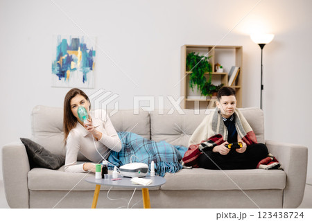 Young woman holding a mask nebulizer and inhaling fumes medication into lungs. Sick lady inhaling through inhaler mask at home and funny boy with gamepad playing video game at home near her Young woman holding a mask nebulizer and inhaling fumes medication into lungs. Sick lady inhaling through inhaler mask at home and funny boy with gamepad playing video game at home near her 123438724