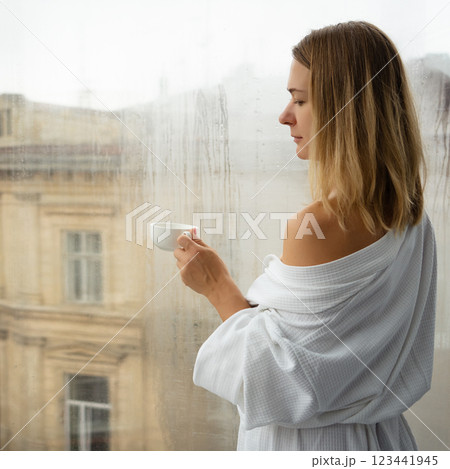 Woman in elegant robe drinking coffee in hotel room and standing near window Woman in elegant robe drinking coffee in hotel room and standing near window 123441945