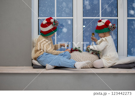 Little children brother and sister in Christmas hats play while sitting on a window with Christmas decor. Little children brother and sister in Christmas hats play while sitting on a window with Christmas decor. 123442273