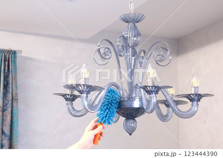 Woman wipe dust on glass chandelier with fluffy broom in living room. Cleanliness and order.  123444390