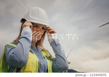 A professional female engineer is meticulously adjusting her safety gear while working on a large wind farm, ensuring both her safety and efficiency in her role within the renewable energy sector 123444970
