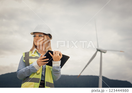 A dedicated female engineer is passionately working on a wind farm project, using advanced machinery in a sustainable landscape and efficiently monitoring the turbines with her tablet 123444980