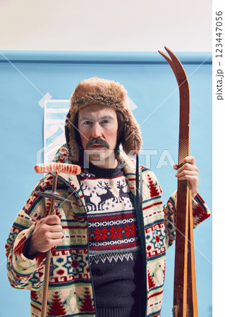 Portrait of man with red cheeks after cold weather wearing fur hat, sweater and textured jacket, holding skis and sausage Portrait of man with red cheeks after cold weather wearing fur hat, sweater and textured jacket, holding skis and sausage 123447056