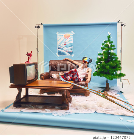 Man lounging on brown leather couch in plaid pajama pants with ski goggles and drink helmet, watching old TV, creating humorous contrast 123447073
