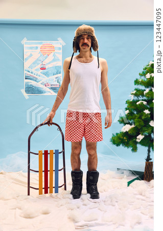 Man wearing white tank top, underwear, fur hat and boots standing with sleds against abstract winter background with artificial snow and tree. Man wearing white tank top, underwear, fur hat and boots standing with sleds against abstract winter background with artificial snow and tree. 123447095