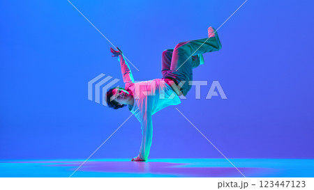Skilled young male dancer captured in mid-motion, showing breakdance technique against gradient blue purple background in neon light Skilled young male dancer captured in mid-motion, showing breakdance technique against gradient blue purple background in neon light 123447123