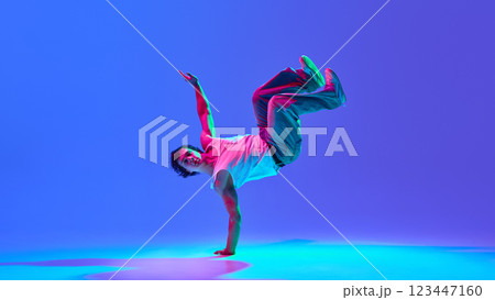 Energetic young man performing breakdance, showing strength and agility against vibrant blue-purple background in neon light. Energetic young man performing breakdance, showing strength and agility against vibrant blue-purple background in neon light. 123447160