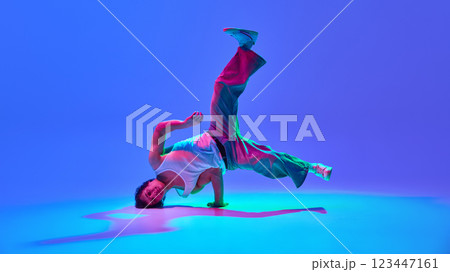 Talented and energetic young man, dancer in motion, performing breakdance against gradient blue purple background in neon light 123447161