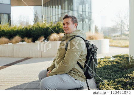 Handsome hipster man model sitting on a bench in the city. Urban lifestyle concept. Traveler. Spring time. Portrait of happy young man in casual clothing looking at camera outdoor. Handsome hipster man model sitting on a bench in the city. Urban lifestyle concept. Traveler. Spring time. Portrait of happy young man in casual clothing looking at camera outdoor. 123451070