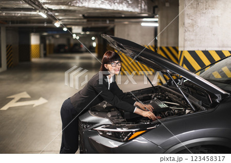 Caucasian businesswoman in formal attire fixes car engine in dimly lit parking garage, conveys independence and problem-solving skills. Caucasian businesswoman in formal attire fixes car engine in dimly lit parking garage, conveys independence and problem-solving skills. 123458317