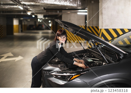 Caucasian businesswoman in formal attire fixes car engine in dimly lit parking garage, conveys independence and problem-solving skills. 123458318