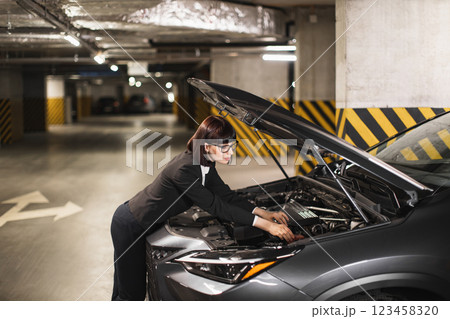 Caucasian adult businesswoman in professional attire repairs car engine under hood in underground parking lot, showcasing independence and problem-solving. Caucasian adult businesswoman in professional attire repairs car engine under hood in underground parking lot, showcasing independence and problem-solving. 123458320
