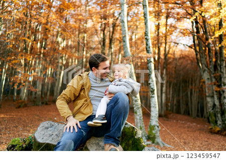 Smiling dad with a little girl on his knees sits on a stump  123459547