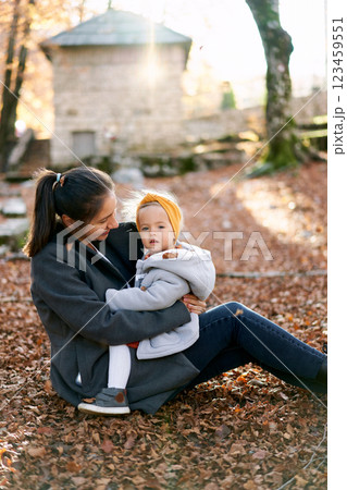 Mom looks at a little girl on her knees, sitting on yellow leaves in an autumn park Mom looks at a little girl on her knees, sitting on yellow leaves in an autumn park 123459551