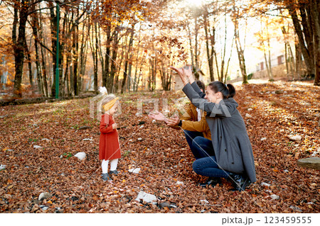 Little girl stands in front of mom and dad squatting and throwing up leaves in the autumn park 123459555