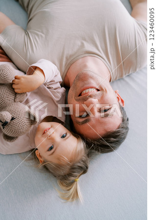 Smiling dad with a little girl and a teddy bear lie on the bed touching their heads. Top view 123460095