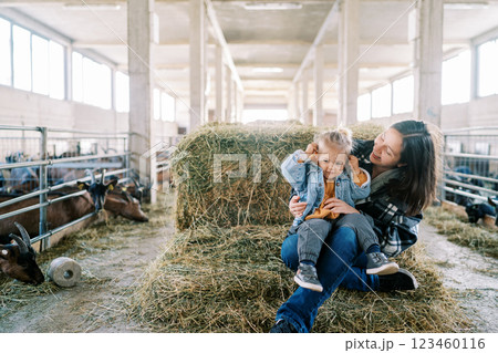 Smiling mom with little girl on her lap sitting on haystack near goat pens at farm Smiling mom with little girl on her lap sitting on haystack near goat pens at farm 123460116