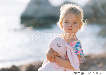Little girl stands by the sea hugging a pink toy hare 123460179