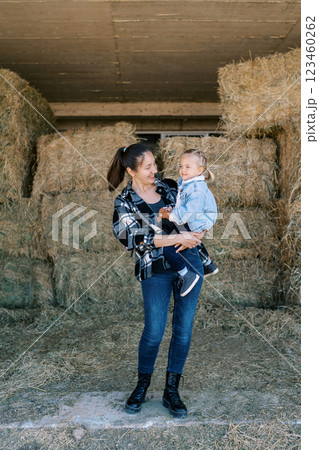 Mom with a little girl in her arms stands near the bales of hay 123460262