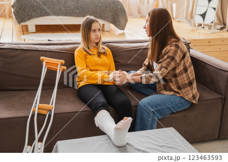 Girl with broken leg in cast sitting with mother on couch at home. Sad teenage kid recovering from broken leg. Girl using cast crutches for quick rehabilitation. Mom supporting daughter Girl with broken leg in cast sitting with mother on couch at home. Sad teenage kid recovering from broken leg. Girl using cast crutches for quick rehabilitation. Mom supporting daughter 123463593