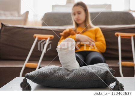 Broken leg. Young girl with broken leg cast sitting on sofa. Sad little kid recovering from broken leg at home. Girl using cast and crutches for rehabilitation. Accident injury fracture treatment 123463637