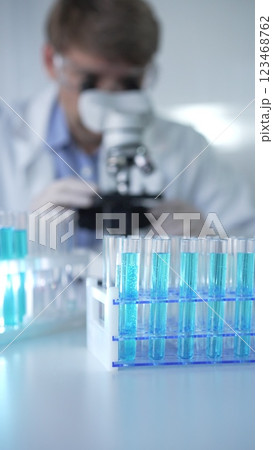 Test tubes with a blue liquid, close-up view. Male scientist wearing white protective gloves and glasses is using microscope on the background. Science and Medicine concept Test tubes with a blue liquid, close-up view. Male scientist wearing white protective gloves and glasses is using microscope on the background. Science and Medicine concept 123468762