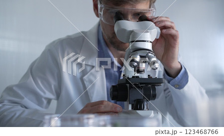 Male scientist wearing lab coat and protective glasses using microscope doing research in modern laboratory setup, portrait view. Low key lighting photo. Science and Medicine concept Male scientist wearing lab coat and protective glasses using microscope doing research in modern laboratory setup, portrait view. Low key lighting photo. Science and Medicine concept 123468766