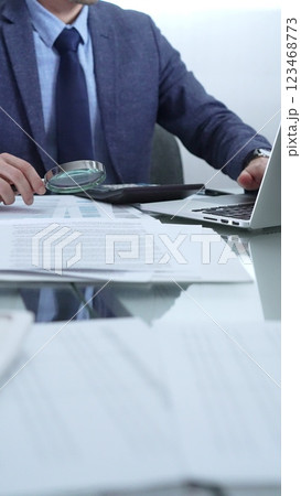 Businessman wearing blue suit is analyzing financial reports with magnifying glass and using calculator in a professional office setting. Low lighting, close-up vertical view. Business people, audit Businessman wearing blue suit is analyzing financial reports with magnifying glass and using calculator in a professional office setting. Low lighting, close-up vertical view. Business people, audit 123468773