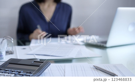 Black calculator, pen and documents are lying on accountant's desk. Businesswoman wearing blue dress is working with documents on the background. Business and audit concept Black calculator, pen and documents are lying on accountant's desk. Businesswoman wearing blue dress is working with documents on the background. Business and audit concept 123468833