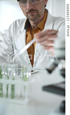 Man science researcher, wearing a lab coat and protective glasses, drips liquid from a pipette into a test tube with a green plant. His face is focused, captured in a vertical portrait. Science and 123468909