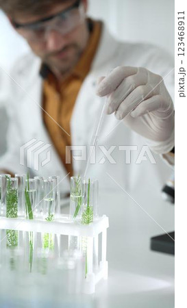 Man scientist wearing a lab coat, white gloves and protective glasses, is dripping liquid from a pipette into a test tube with a green plant inside, vertical close up view. Science and medicine 123468911