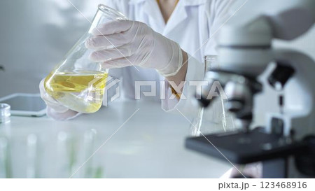 Microscope is near a scientist, wearing a lab coat and white protective gloves, and holding beaker with a yellow oily liquid in laboratory setting, close up. Medicine and science concept Microscope is near a scientist, wearing a lab coat and white protective gloves, and holding beaker with a yellow oily liquid in laboratory setting, close up. Medicine and science concept 123468916