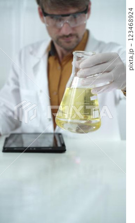 Man scientist wearing white protective gloves, and glasses is holding a yellow chemical solution inside an Erlenmeyer flask in a laboratory, vertical portrait 123468924