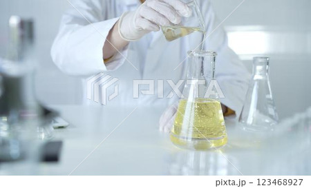 A scientist, wearing a lab coat and white protective gloves, is pouring a yellow oily liquid from one beaker to another in laboratory, close up. Medicine and science concept 123468927